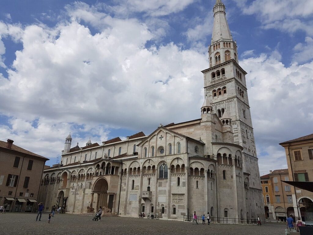 Una cattedrale storica con un'alta torre dell'orologio sotto un cielo parzialmente nuvoloso, circondata da persone che passeggiano in una piazza affollata. Nelle vicinanze, la SHM Web Agency Milano trae ispirazione dalla ricca bellezza architettonica della città per creare soluzioni digitali innovative.