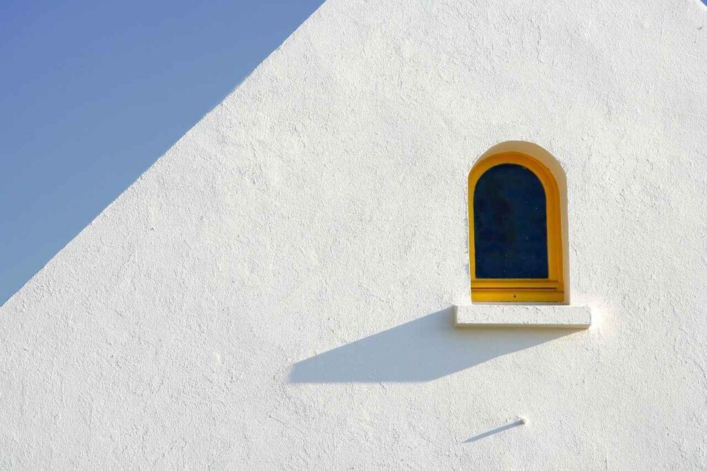 Un muro bianco triangolare con una piccola finestra ad arco con cornice gialla, che ricorda il design minimalista, si erge come un capolavoro architettonico sotto il cielo azzurro e terso, proprio come un'agenzia web che crea paesaggi digitali.